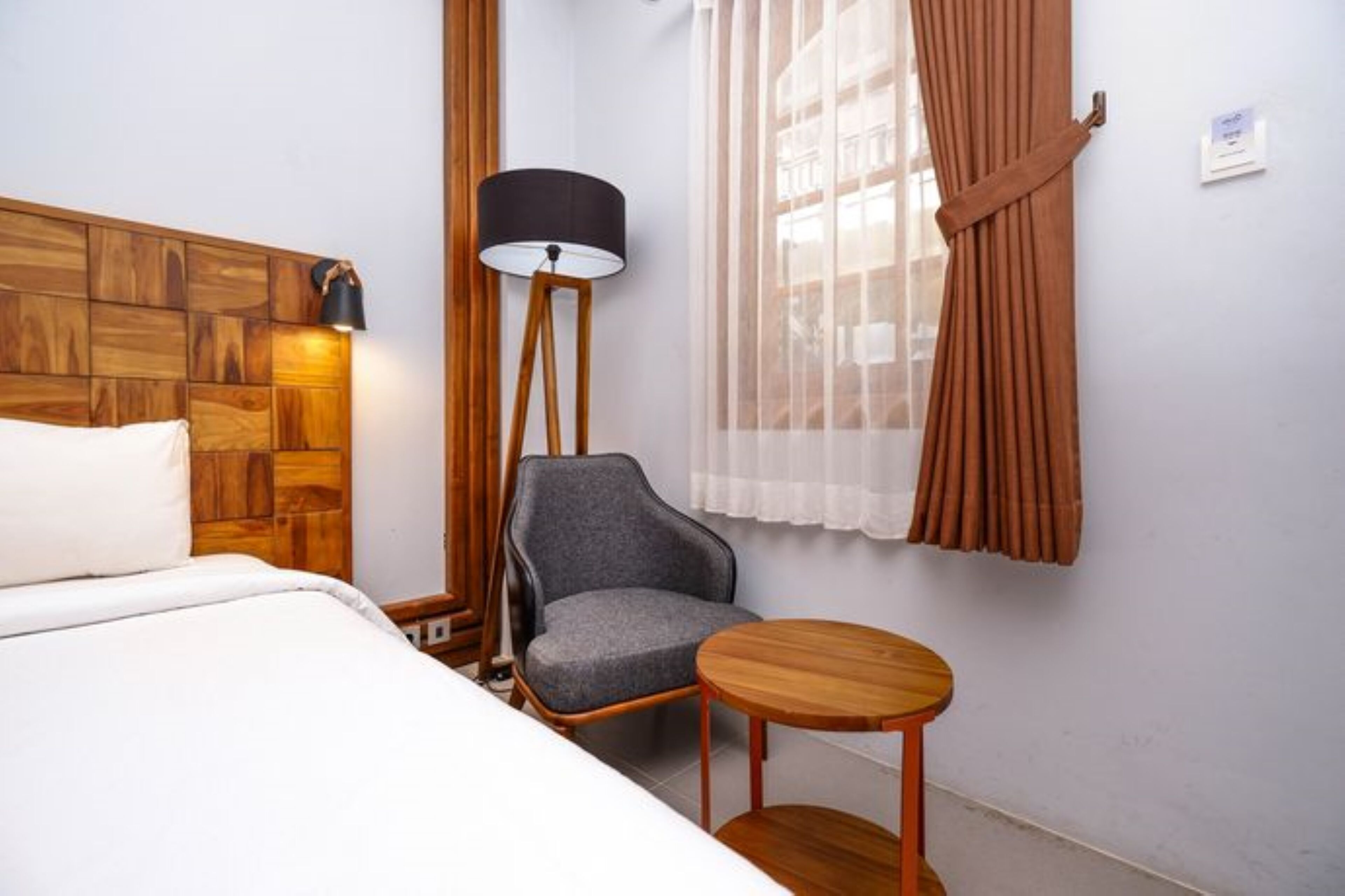 A hotel room with a wooden headboard, a grey armchair, a small wooden table, a floor lamp, and a window with sheer curtains.