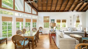 A spacious living room with a wooden ceiling, large windows with bamboo blinds, a glass dining table, wicker chairs, a grey sofa, and a wooden coffee table.
