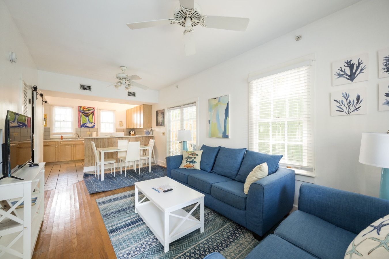 A modern living room with a blue sofa, white coffee table, and a TV. There is a kitchen in the background with wooden cabinets and a dining area.