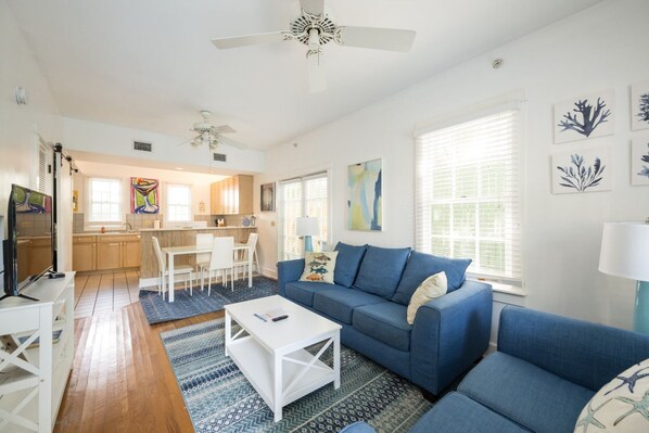 A modern living room with a blue sofa, white coffee table, and a TV. There is a kitchen in the background with wooden cabinets and a dining area. - Coco`s Poolside Retreat (Key West)