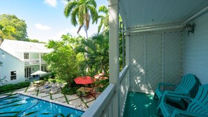 A pool area with lounge chairs and a red umbrella, surrounded by lush greenery and a white building.