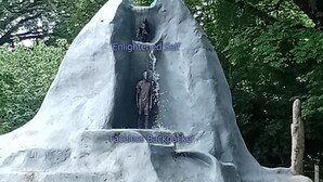 A large, abstract sculpture resembling a mountain with a waterfall feature, set in a park with trees in the background. - Backpackers Temple  (Mae Sot)