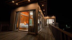 Una habitación de hotel moderna con techo de madera, un amplio ventanal corredero que da a un balcón y una cama con ropa de cama blanca.