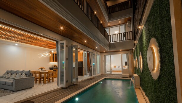Un área de piscina interior moderna con cubierta de madera, un sofá gris y un comedor.