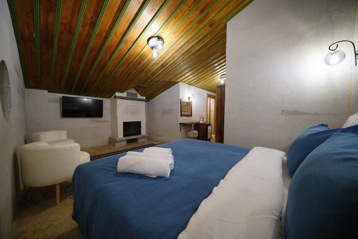 Un dormitorio con techo inclinado, chimenea, televisor y cama con ropa de cama azul y blanca.