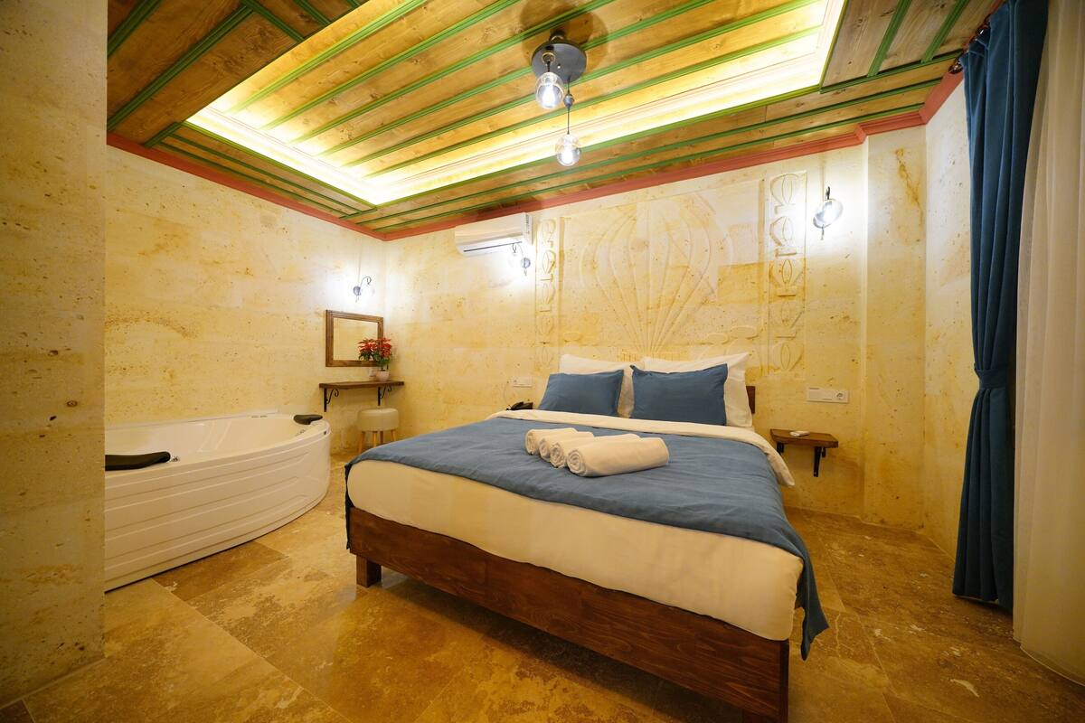 Un dormitorio con techo de madera, una cama grande con ropa de cama azul, una bañera blanca y una mesita de noche pequeña con lámpara.