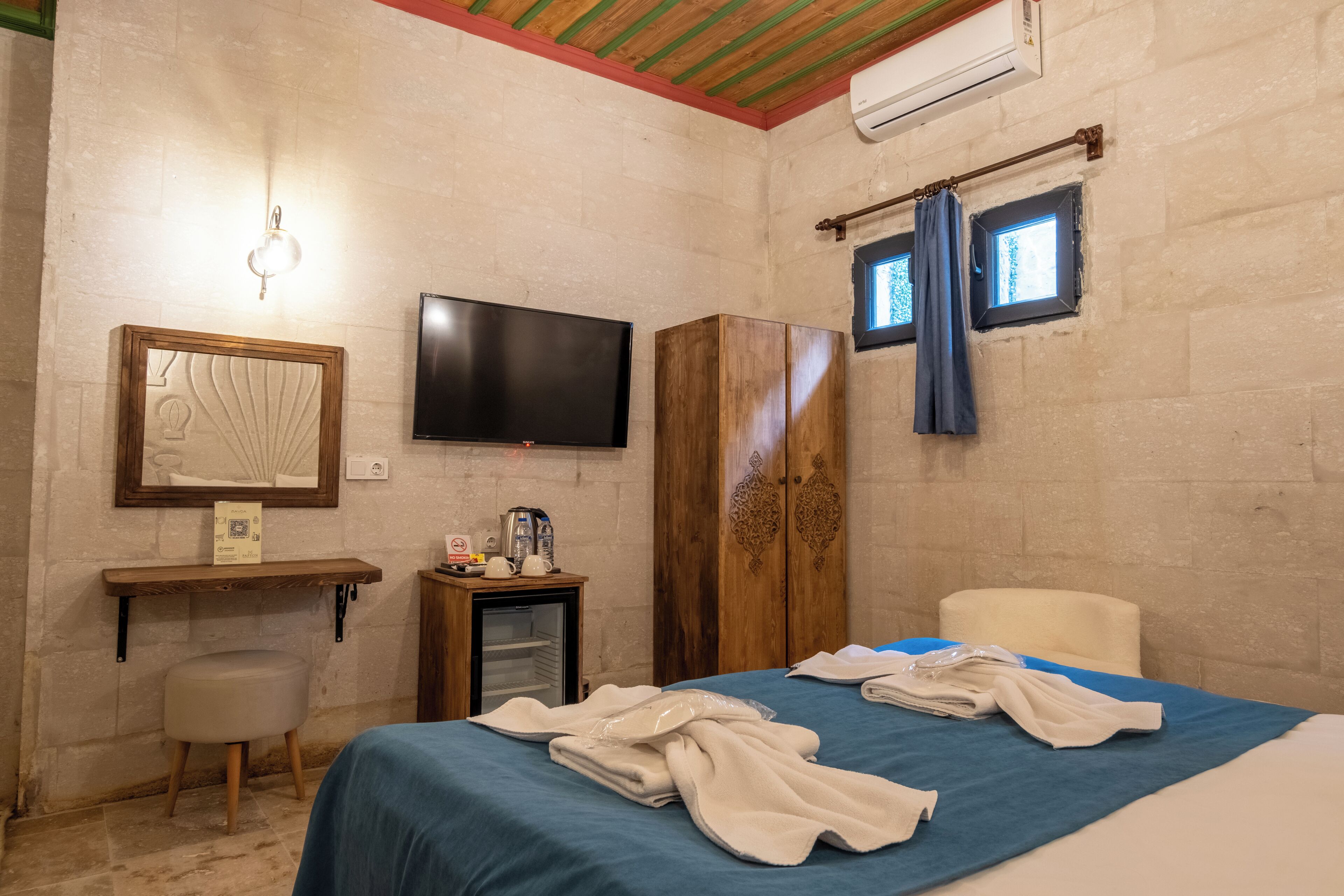 A hotel room with a bed, a television, a wardrobe, a small refrigerator, and a window with curtains.