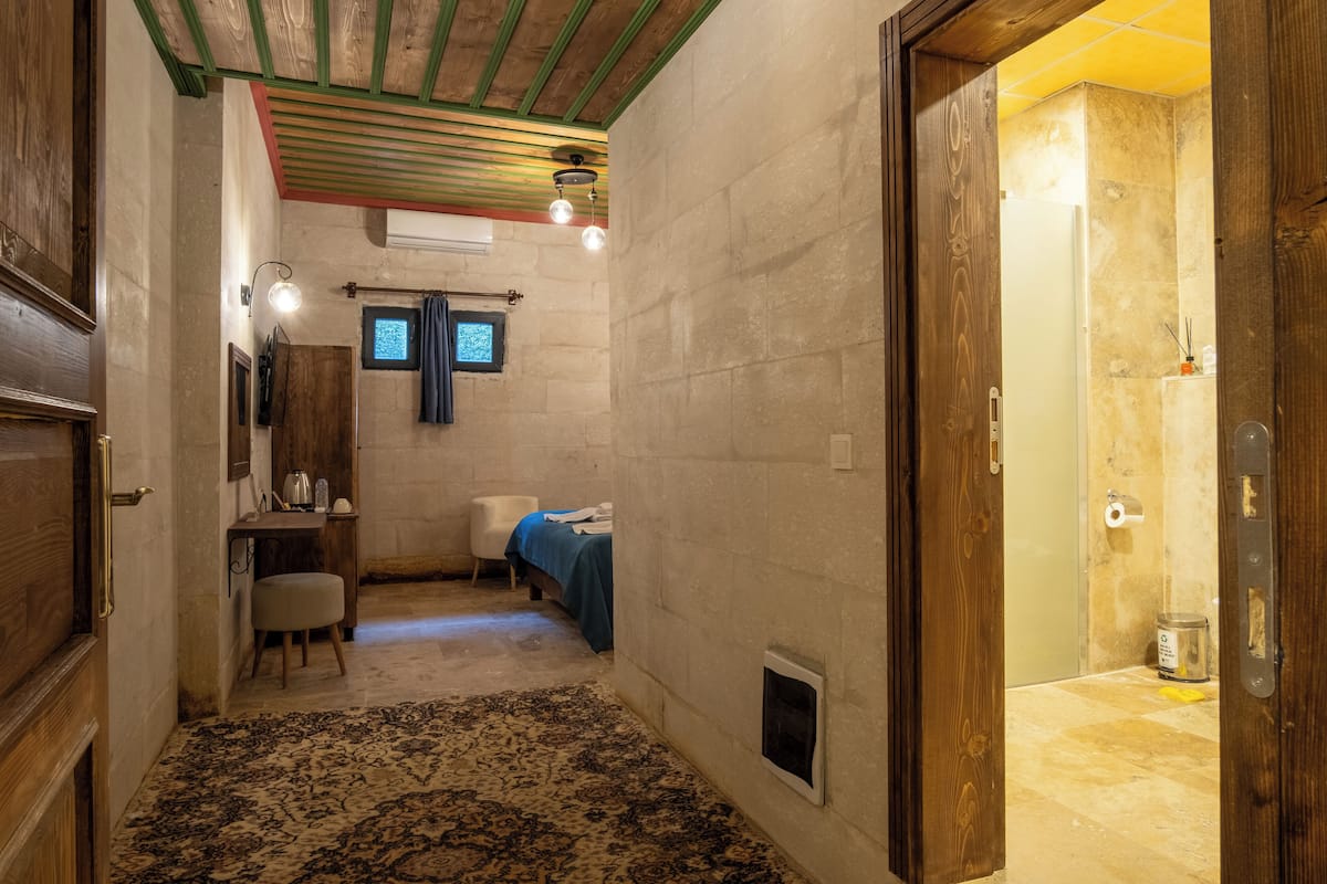 Habitación pequeña con cama, escritorio y silla. Tiene una ventana con cortinas, un radiador y una puerta que da a otra habitación.