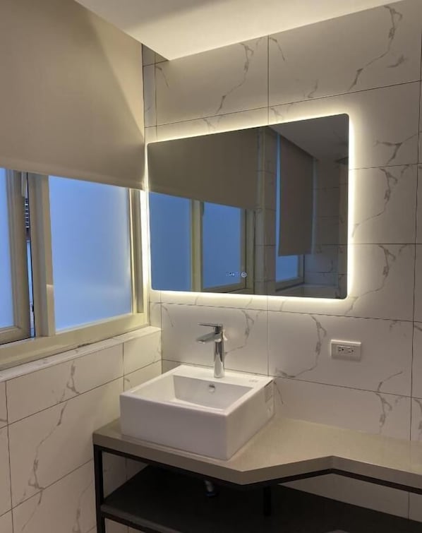 Un bagno moderno con pannello di marmo bianco, uno specchio rettangolare con illuminazione integrata, un lavabo a colonna bianco e una finestra con tende.