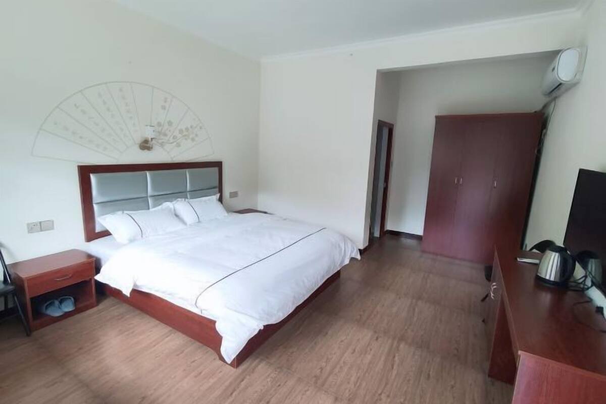 A hotel room with a bed, a wooden nightstand, a desk, a wardrobe, and a wall-mounted fan.