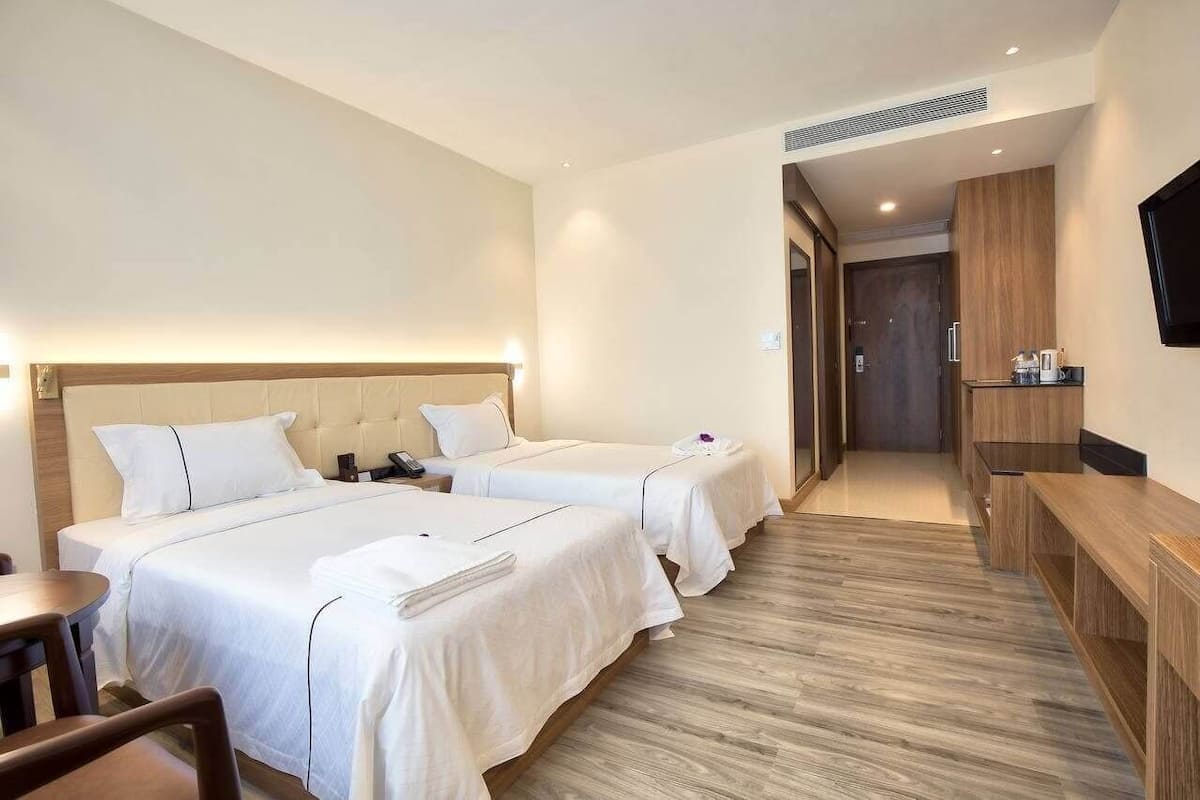 A hotel room with two beds, a wooden floor, a TV mounted on the wall, and a small table with a chair.