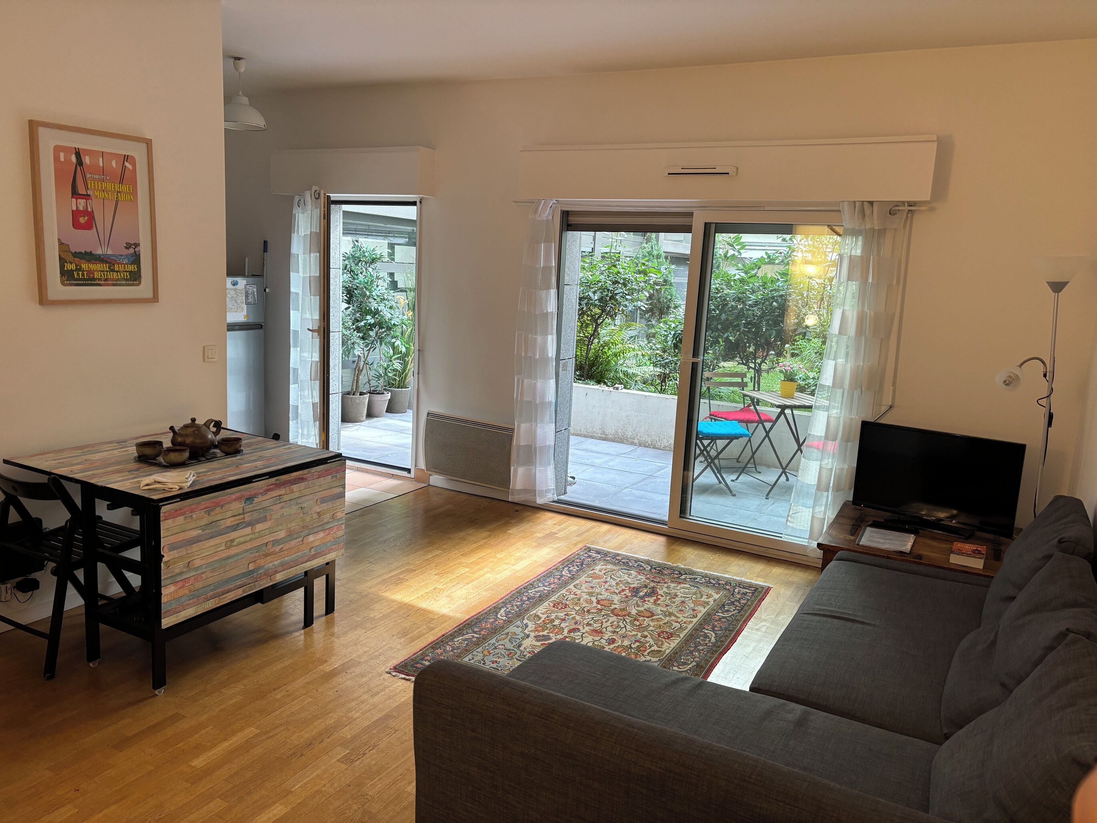A living room with a sofa, a TV, a dining table with a teapot, and a view of a balcony with plants and a table.