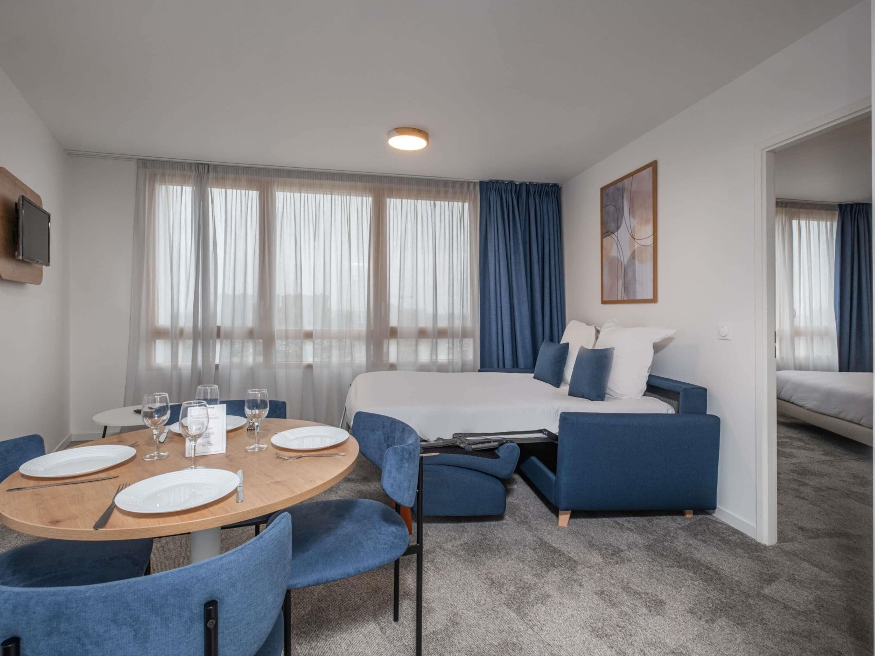 Une chambre d’hôtel avec un lit, une table à manger avec des chaises, un téléviseur fixé au mur et une grande fenêtre avec des rideaux.
