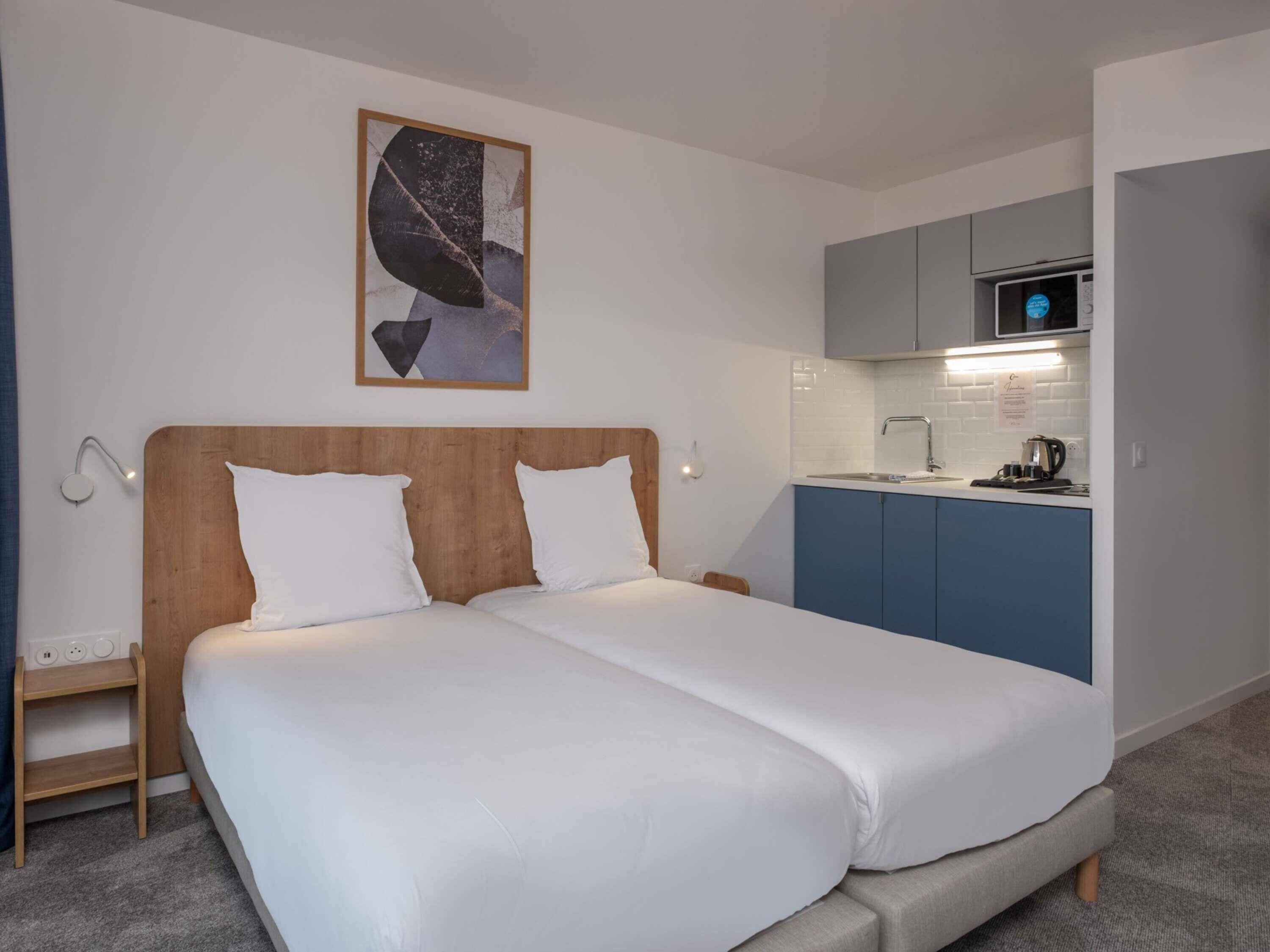 A double bed with white linens, a wooden headboard, a small bedside table, a wall-mounted lamp, a kitchen area with a microwave, and a blue cabinet.