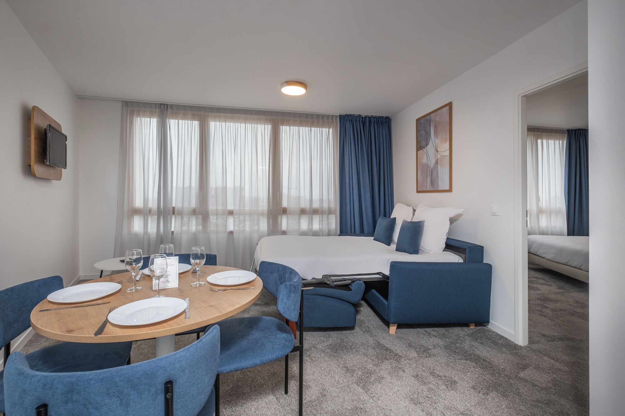 Una habitación de hotel moderna con una mesa de comedor redonda, sillas tapizadas en azul, una cama con ropa de cama azul y un gran ventanal con cortinas translúcidas.