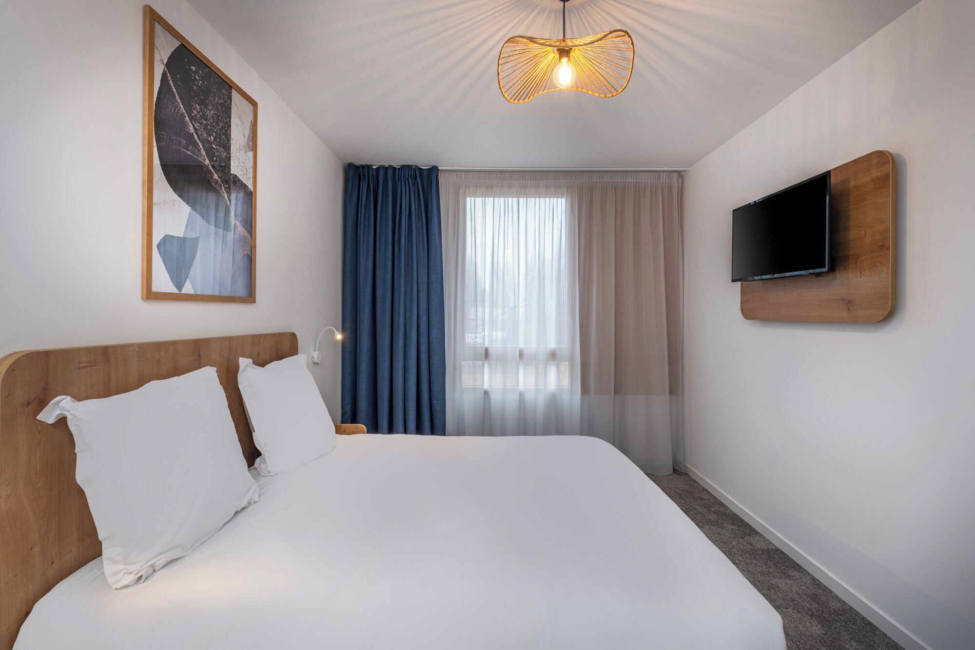 A hotel room with a bed, a TV mounted on the wall, a ceiling light fixture, and a window with curtains.