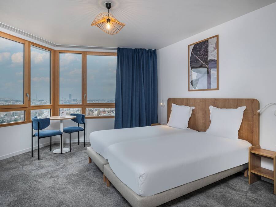 A hotel room with a large bed, a desk, two chairs, a view of the city, and a ceiling light.