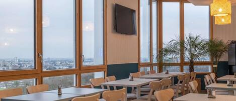 A restaurant with multiple tables and chairs, large windows offering a city view, and a television mounted on the wall.