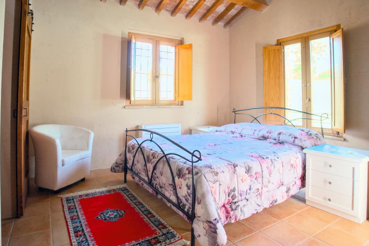 Une chambre à coucher comprenant un lit, une chaise, une table de chevet et une commode. Deux fenêtres laissent entrer la lumière naturelle.