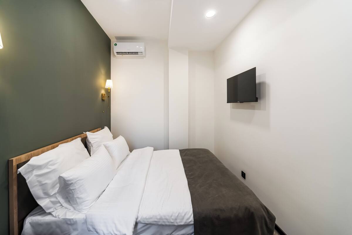 Una cama bien hecha con ropa de cama blanca y gris, un televisor de pantalla plana montado en la pared y un aire acondicionado también instalado en la pared.