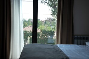 A hotel room with a bed, curtains, and a view of buildings and trees outside. - Estetico Boutique Hotel (Yerevan)