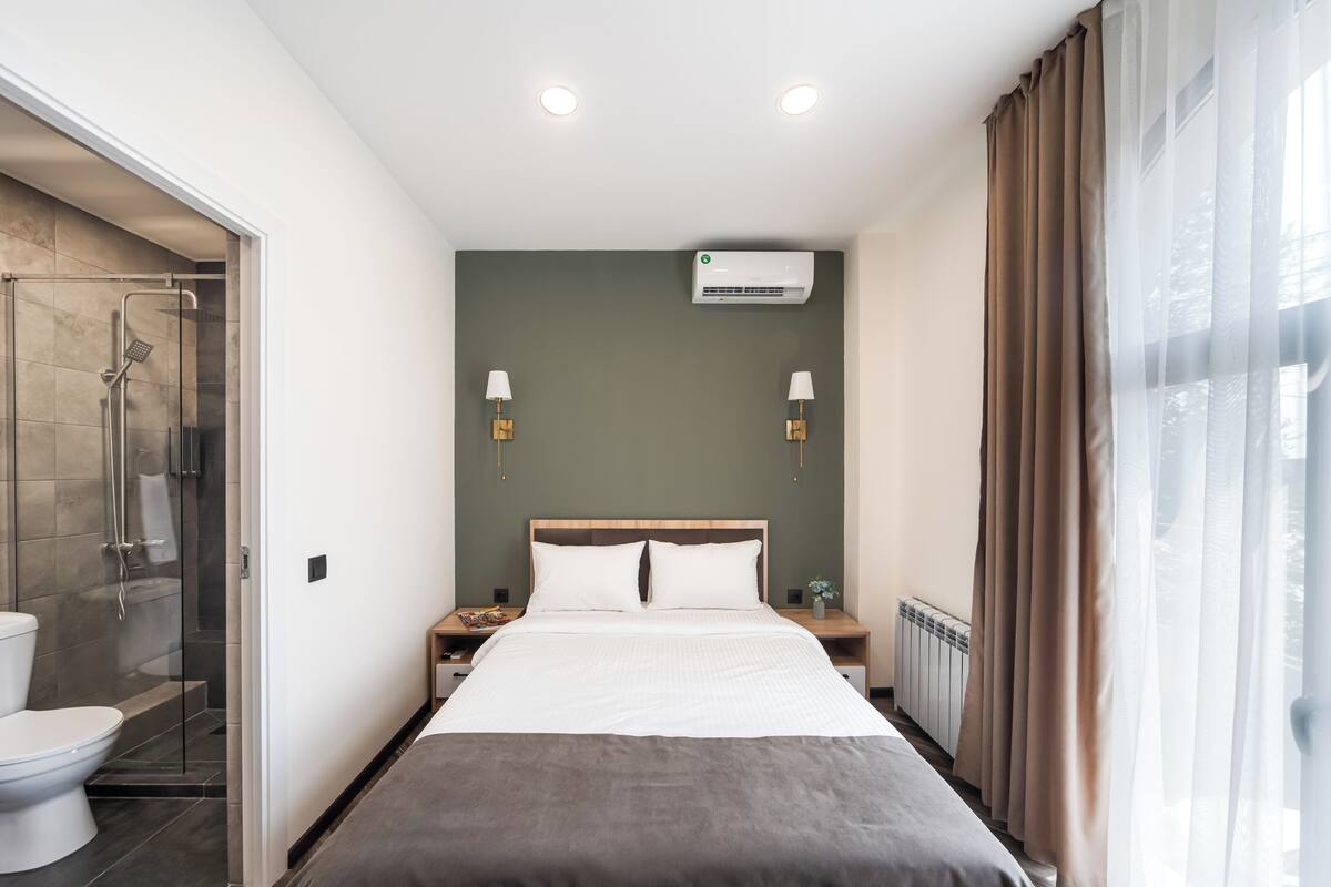 Habitación de hotel moderna con una cama grande, ducha, inodoro y ventana con cortinas.