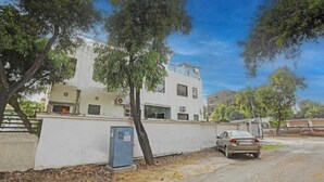 Un edificio de dos pisos con fachada blanca, un coche estacionado al frente y árboles al fondo.