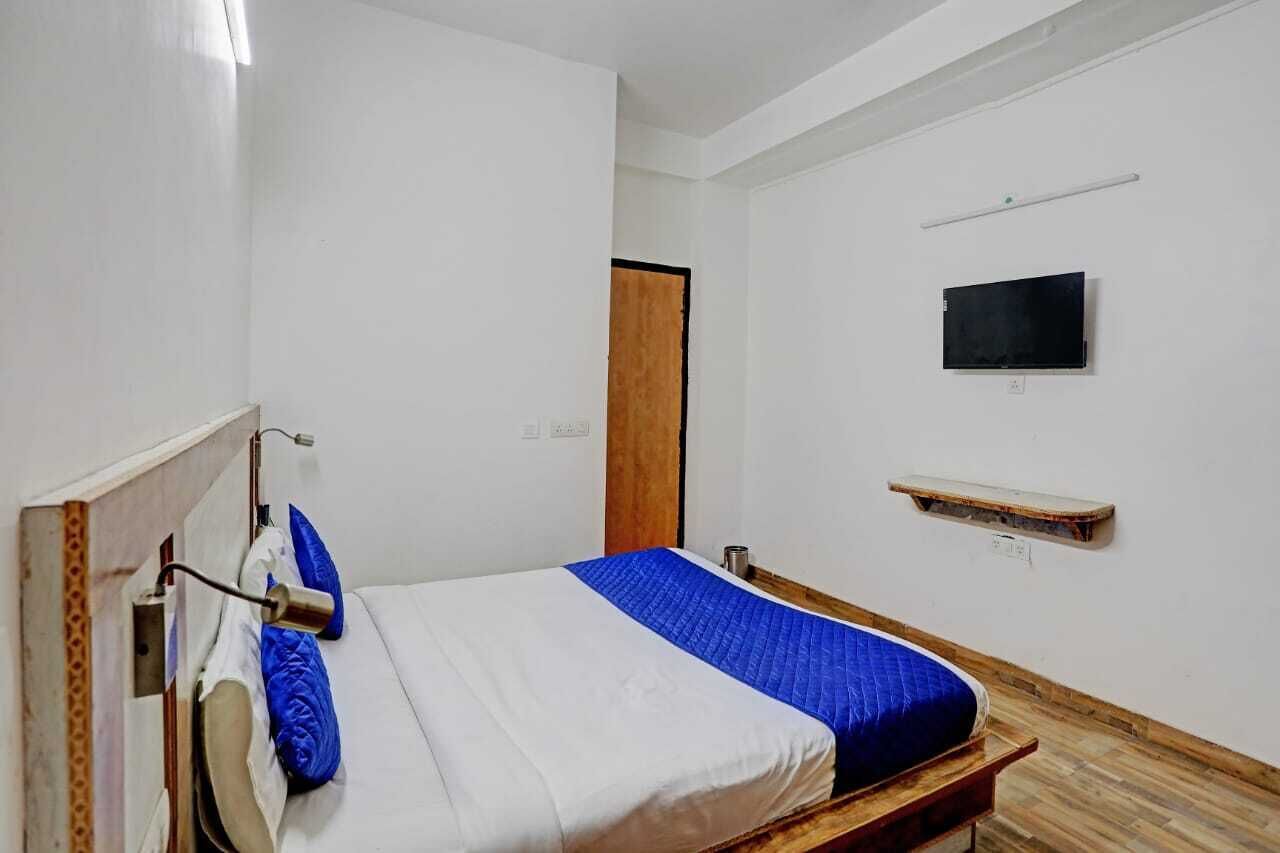 Camera con un letto singolo, una testiera in legno, una coperta blu e bianca, una televisione a parete e uno scaffale in legno.