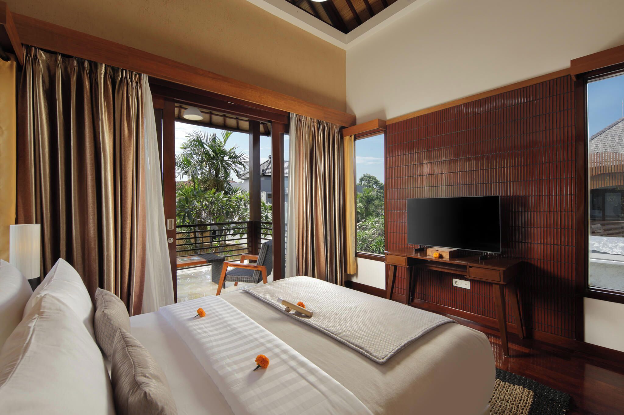 A hotel room with a bed, a TV, a balcony with a chair, and a view of greenery.
