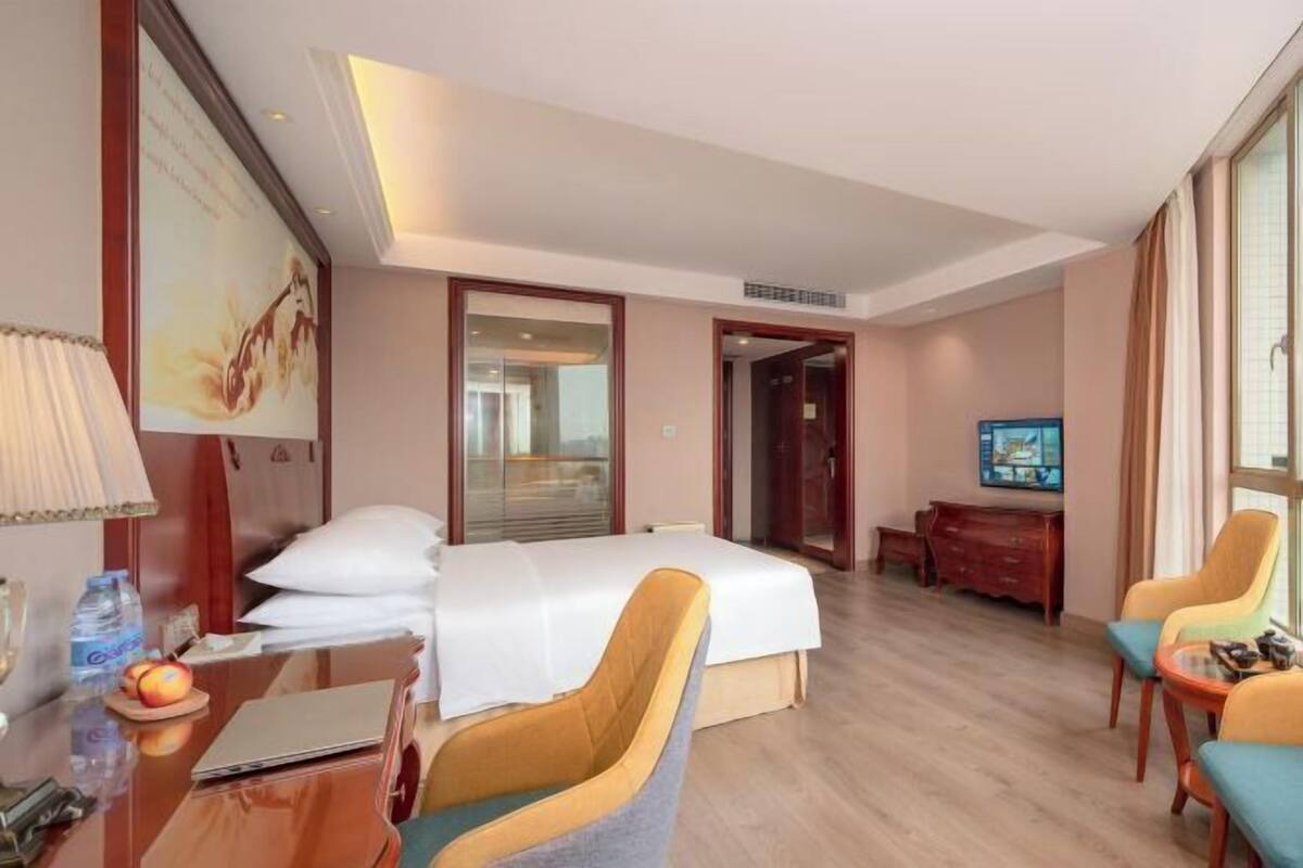 A hotel room with a bed, a desk with a laptop, a chair, a television, and a large window.