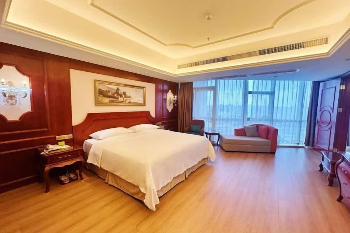 A hotel room with a large bed, a desk, a chair, a sofa, and a large window with curtains.