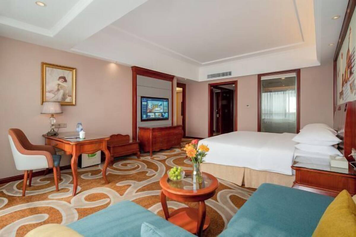 A hotel room with a bed, a desk, a chair, a TV, and a small table with flowers.