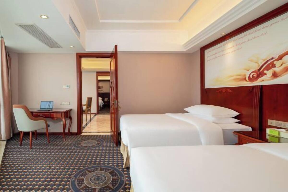 A hotel room with two beds, a desk with a laptop, a chair, and a patterned carpet.