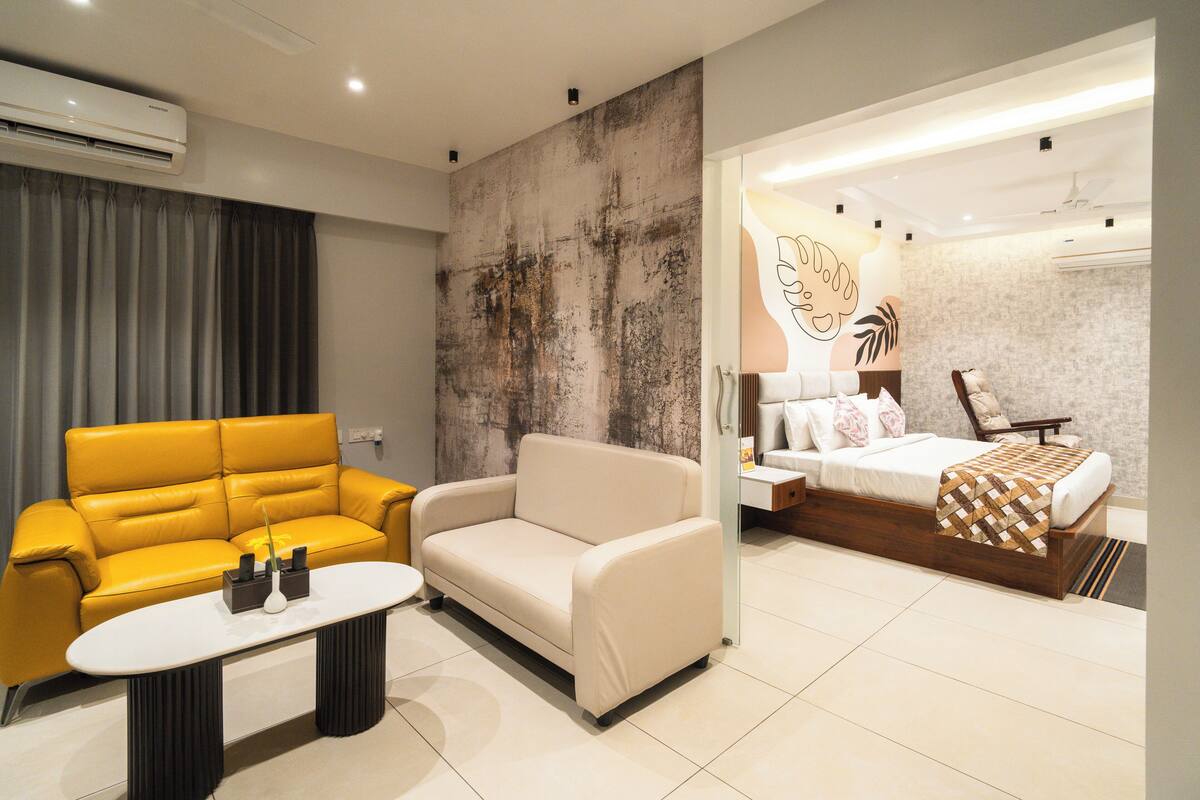 Ein modernes Hotelzimmer mit einem gelben Sofa, einem beige gefärbten Sessel, einem kleinen weißen Tisch und einem Bett mit Holzrahmen.