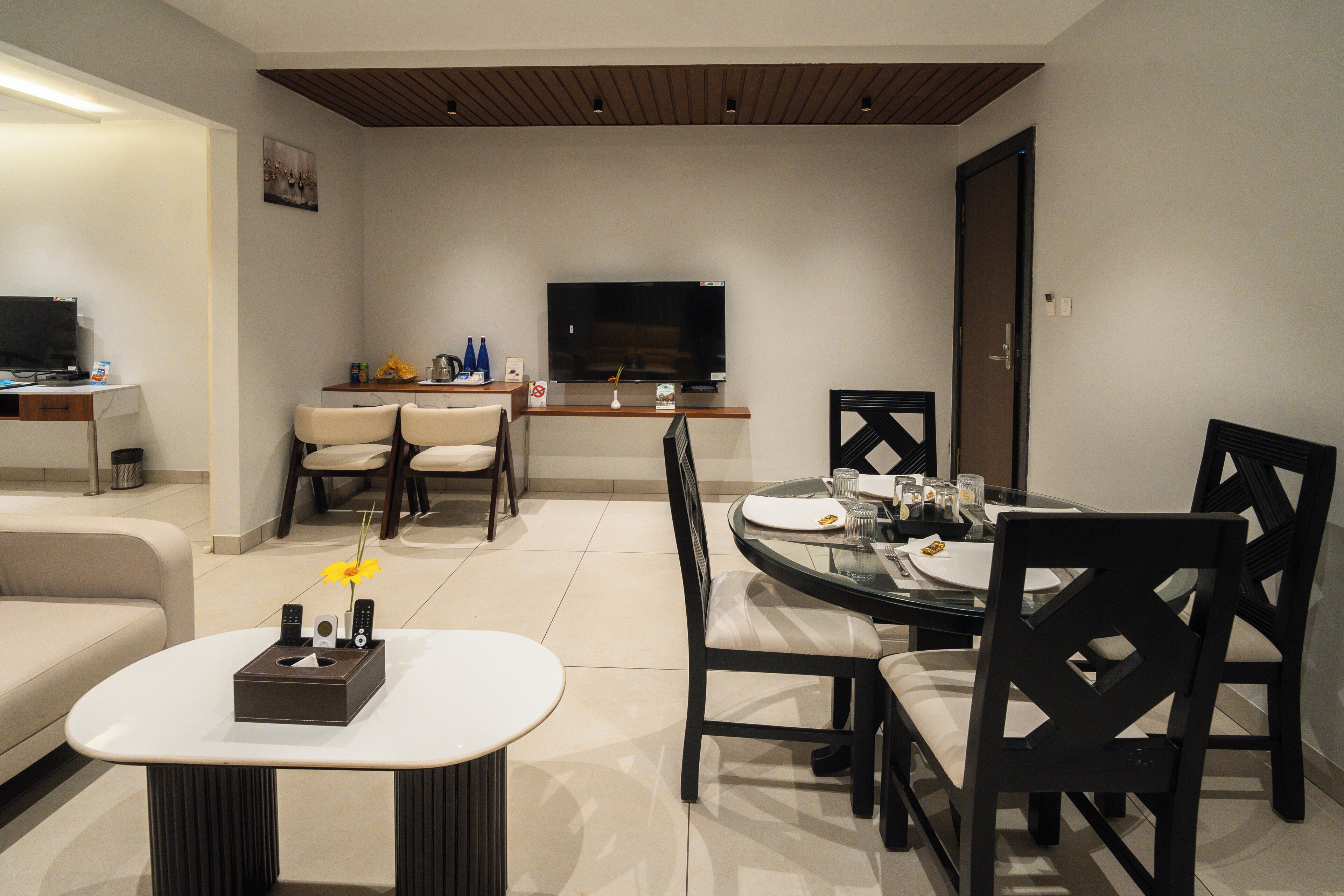 A modern dining area with a round glass table, black chairs, and a white sofa. A flat-screen TV is mounted on the wall above the table.