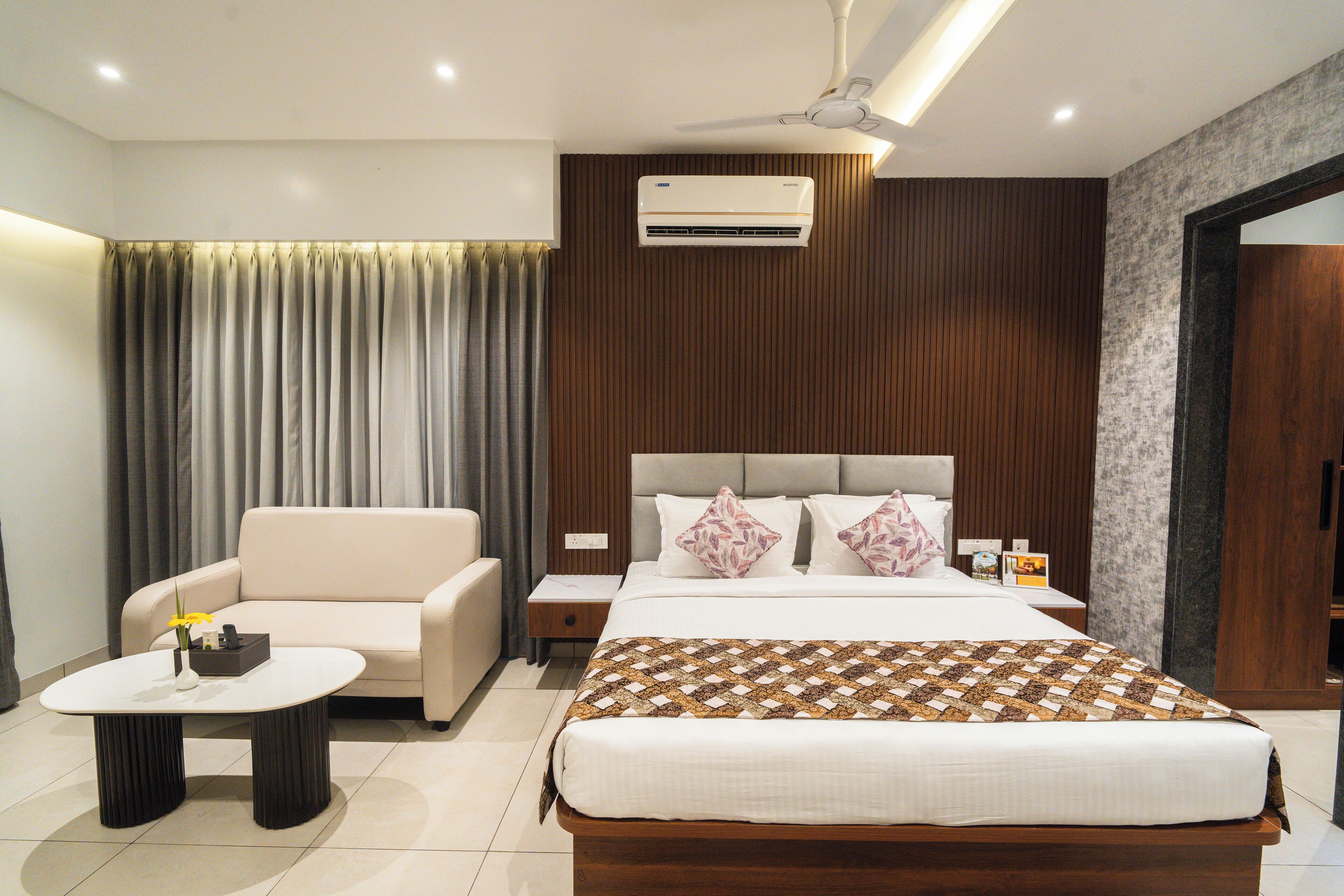 A modern hotel room with a bed, a sofa, a small table, and an air conditioning unit.