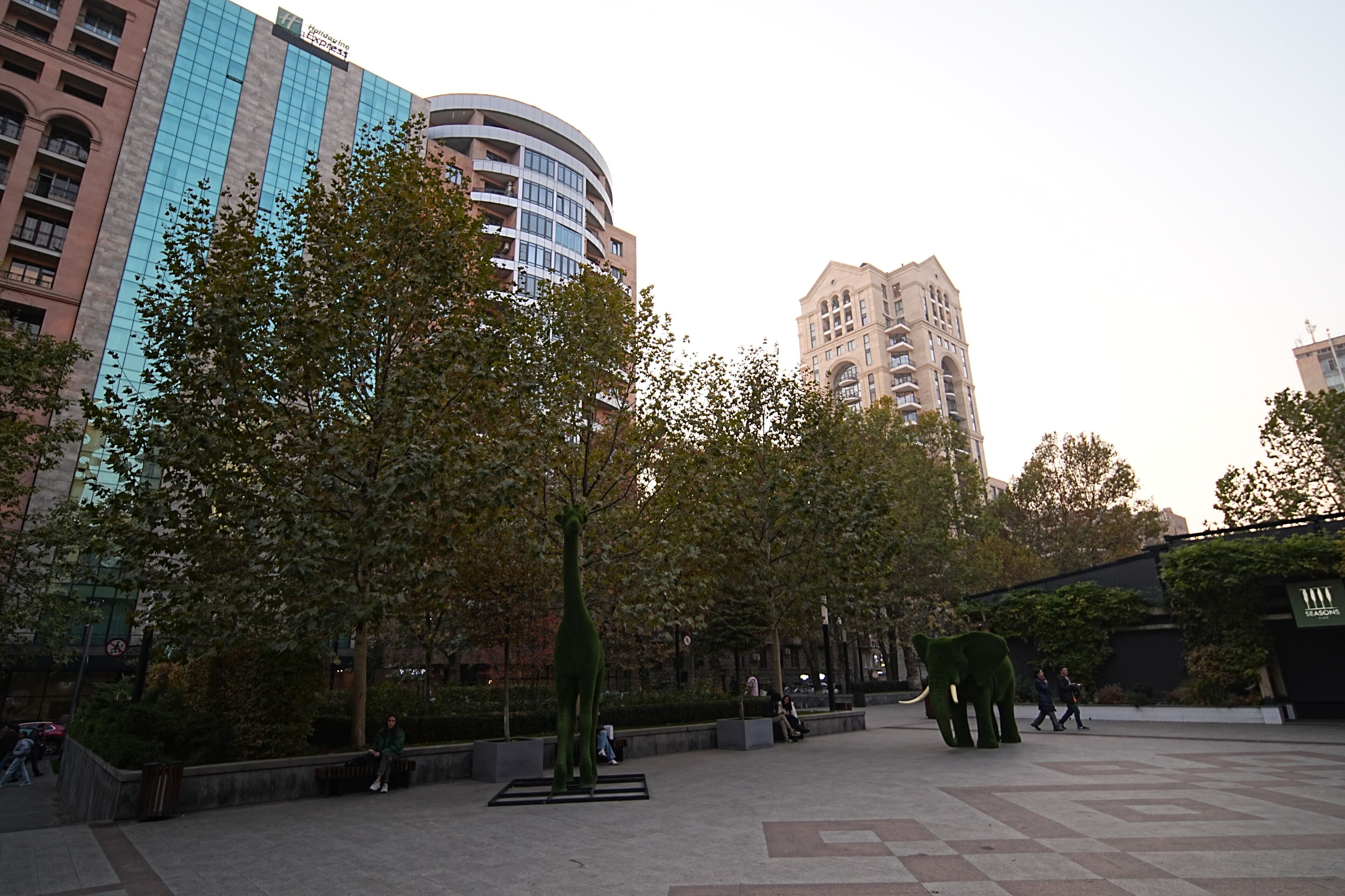 A plaza with modern buildings, a giraffe sculpture, and people walking.