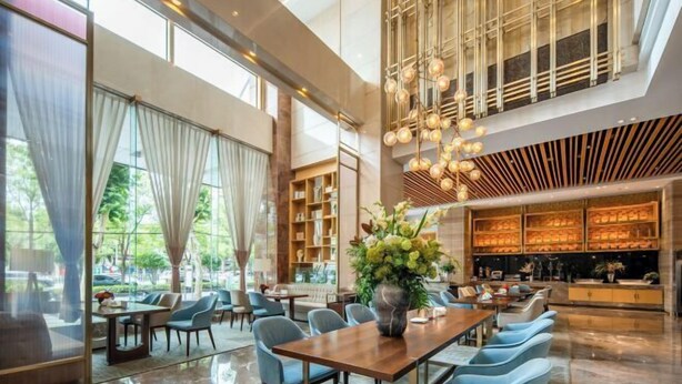 A spacious hotel lobby with a high ceiling, large windows, and a central seating area with a wooden table and chairs.