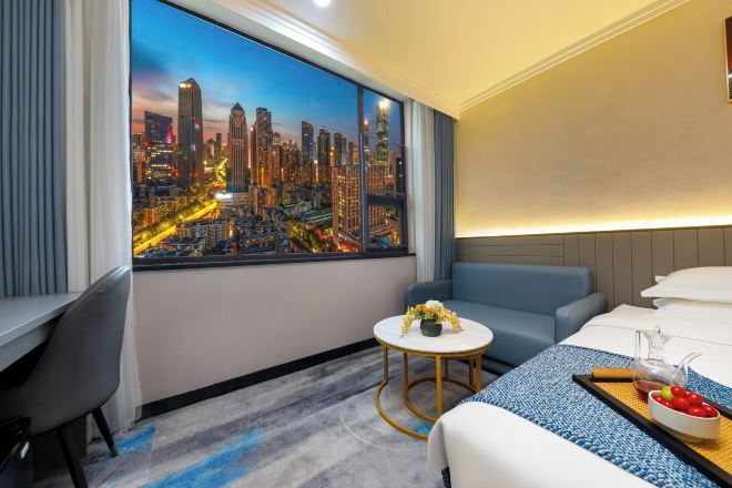 A hotel room with a large cityscape view, a bed, a small round table with a vase of flowers, and a blue sofa.