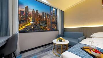 A hotel room with a large cityscape view, a bed, a small round table with a vase of flowers, and a blue sofa.