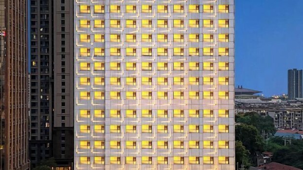 Un edificio alto con una cuadrícula de ventanas uniformemente iluminadas, rodeado de otras estructuras elevadas.