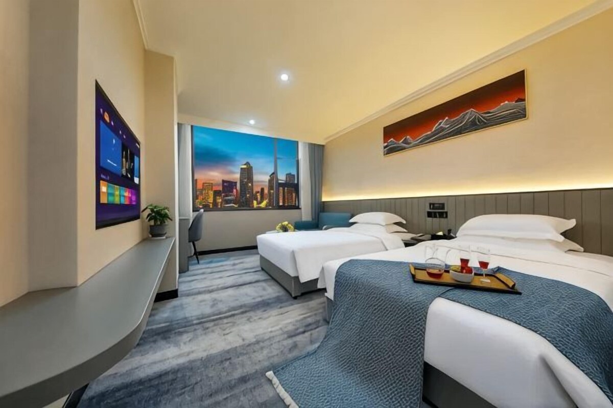 A hotel room with two beds, a cityscape painting, a digital clock, and a tray with cups and a bottle.