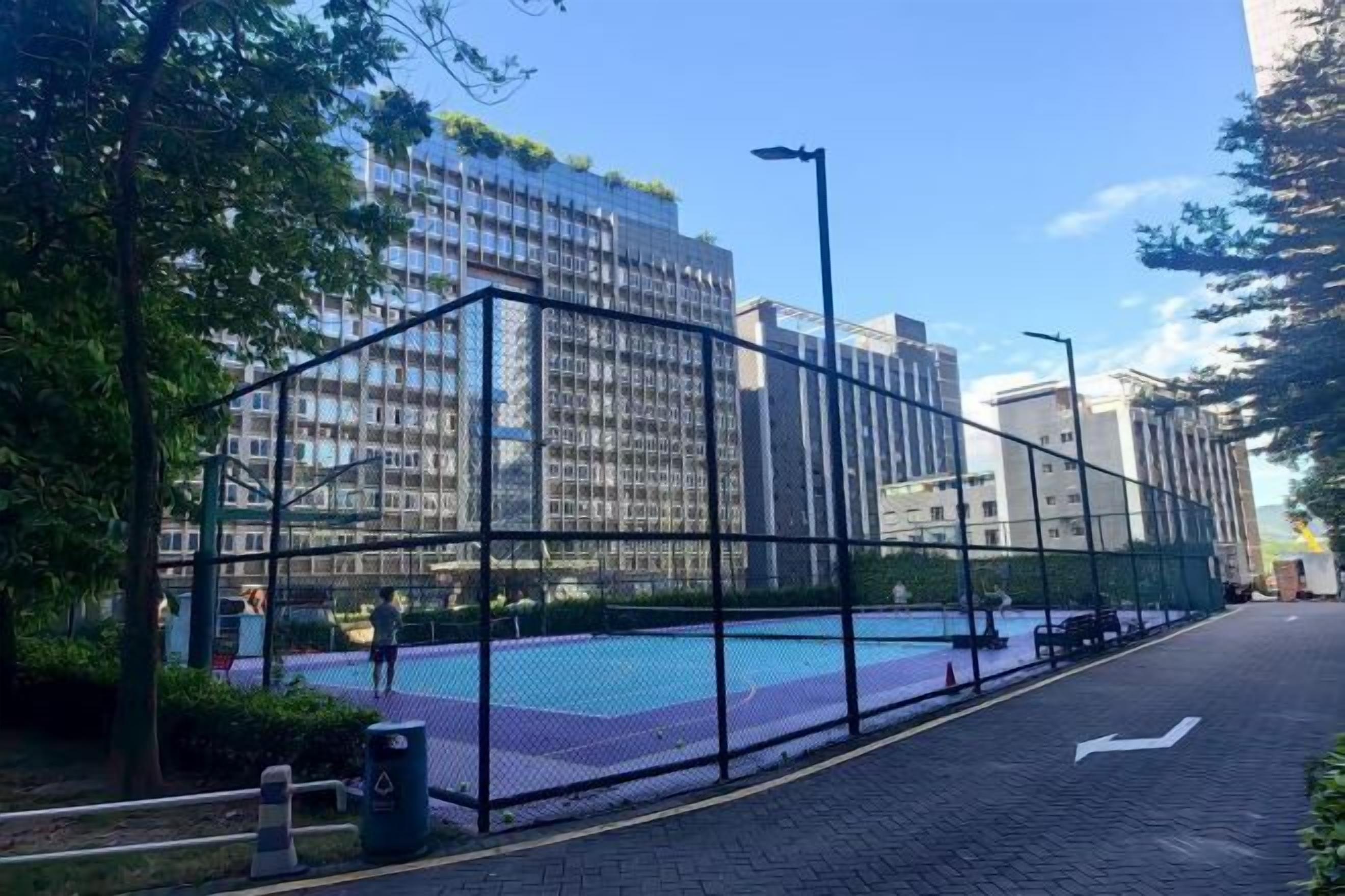 A fenced outdoor area with a basketball court, surrounded by tall buildings and trees.