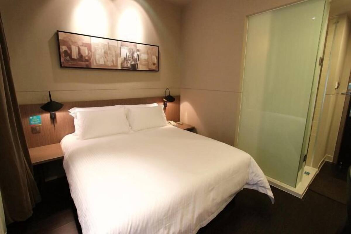 Une chambre d’hôtel comprenant un lit, une table de chevet, une lampe fixée au mur, une cabine de douche en verre et un tableau encadré accroché au mur.