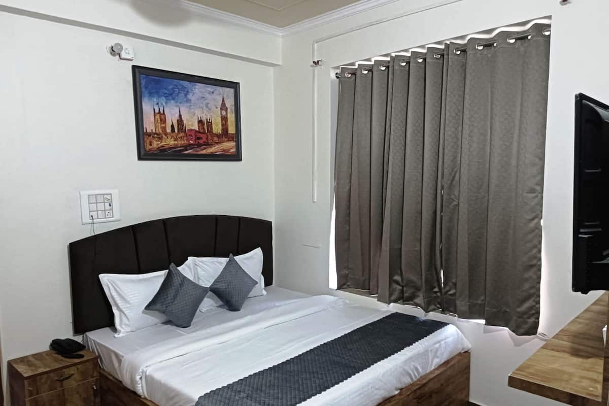 A bedroom with a bed, a nightstand, a wall-mounted power outlet, a painting of a cityscape, and a window with curtains.