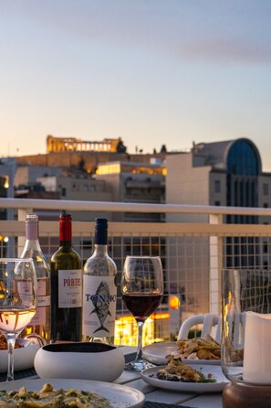 A table set with wine bottles, glasses, and plates of food on a balcony with a cityscape view. - Parthenon View Luxury Penthouse (Athens)