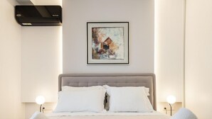 A neatly made bed with white linens and two rolled towels, a framed abstract painting on the wall, and a modern air conditioning unit above the bed. - Parthenon View Luxury Penthouse (Athens)