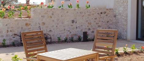 A patio with two wooden chairs and a table, a stone wall, and a row of flowers.