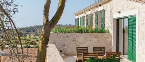 A stone building with green shutters, a patio with wooden furniture, and a view of a vineyard.