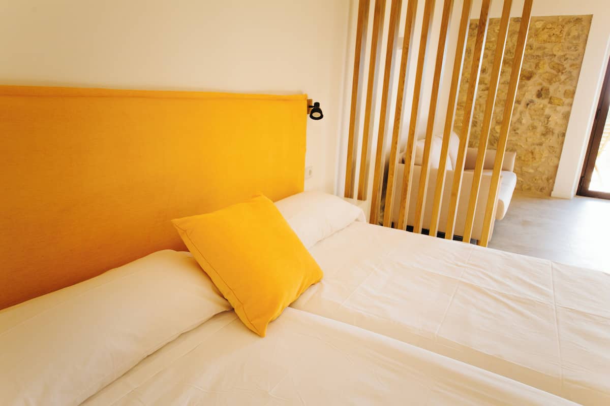 A bed with white bedding and a single yellow pillow, a wooden headboard, and a stone wall in the background.